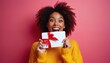 © Pete - Happy young woman with afro hairstyle holds white gift card with red ribbon. She expresses excitement, joy on vibrant pink background. Female in yellow sweater shows gift card, smiling with wide eyes.