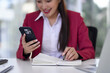 © Pichsakul - Professional Asian Woman Engaging Smartphone While Taking Notes Work Desk Office Environment Modern Business Productivity