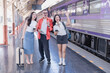 © Tj - Happy friends on a train platform in Thailand, smiling and waving with luggage as they wait for departure, enjoying vacation, conversation and urban travel excitement