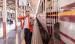 © Tj - Excited adult tourist with backpack and map raising arm in celebration, standing by a waiting train at a bustling railway platform, eagerly anticipating a new adventure