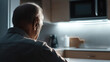 © photo for everything - Thoughtful older adult seated in a softly lit kitchen setting, facing away from the camera. The individual appears contemplative in this quiet, dimly lit scene.
