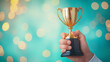 © kampolsuk - Golden trophy held by hand against teal bokeh background celebrating victory and achievement with warm light and festive mood