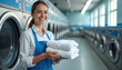 © Viktor - Smiling woman in blue apron holds stack of clean white towels. Pro laundry service employee stands by rows of washing machines and dryers in bright laundromat. She works in clean facility.
