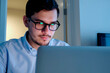 © pressmaster - Portrait of young adult Caucasian man with short dark hair and glasses looking at laptop screen, sitting indoors, focused expression, working or studying on computer