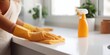 © Xenia_Stash - Woman in yellow gloves wiping a white kitchen counter with an orange microfiber cloth, a spray bottle beside her. Home cleanliness concept.