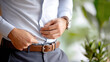 © M - Man adjusting his light blue dress shirt while wearing a brown belt, showcasing a professional and polished appearance in a green setting