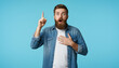 © Viktor  Shmihinskyi - Bearded man in casual shirt pointing up with surprised face, solid cyan blue background.