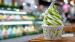 © M - Swirled green and white soft serve ice cream in a cup on a wooden table with blurred dessert display in the background