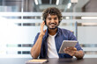 © Tetiana - Portrait of a young Indian man in a headset sitting in an office in front of the camera, holding a tablet and talking to the camera
