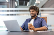 © Tetiana - A young Indian man wearing headphones sits at a desk in the office, working on a laptop and holding a cup of coffee in his hand