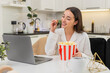© Andrii Iemelianenko - Young woman at home watches trailer on laptop eats popcorn laughs and relaxes fun break. Caucasian girl on sofa sits at table watches video smiles bright mood evening snack casual chill online clip.