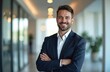 © miss irine - Handsome businessman stands in office. Man wears suit smiling confidently. He is a successful professional with arms crossed. Modern office interior blurred in the background.