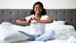© Prostock-studio - A joyful black woman sits cross-legged on her large bed, smiling and making a heart gesture with her hands. She feels happy and relaxed in her cozy bedroom, embracing a wonderful morning.