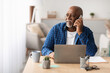 © Prostock-studio - A senior Black man is seated at a table in his office, using a laptop while speaking on a cellphone. He appears engaged in a work conversation, looking aside thoughtfully.