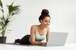 © Prostock-studio - A young woman in athletic clothing lies comfortably on a fitness mat, focused on her laptop. She smiles happily while browsing online, enjoying her time at home with a green plant nearby.