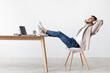 © Prostock-studio - A calm man sits at his desk in a serene setting, wearing wireless headphones and listening to soothing lounge music. He leans back with his legs on the table, smiling as he relaxes.