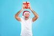 © Prostock-studio - A mature man wearing a red Santa hat joyfully holds a Christmas present above his head in a blue studio. He expresses excitement about the holiday season and gift giving.