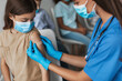 © Prostock-studio - Preteen girl sits in a clinic as a nurse administers a covid-19 vaccine. The nurse carefully applies a medical plaster to the girl's arm following the injection. Other children wait nearby.