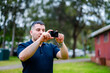 © Austockphoto - A man in his 30s stands in a park, taking photographs with a digital camera on an overcast day