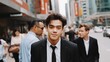 © liliyabatyrova - An Asian man in a classic black suit and tie stands confidently, facing the camera. City buildings tower around him as people stroll by under the blue sky, creating an urban scene full of life