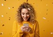 © Vasiliy - Happy excited woman looking at her smartphone under falling confetti. Young girl celebrating a win or receiving good news on a yellow background. Online promotion concept with copy space