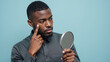 © Jari.art - Man examines his reflection small mirror, showcasing thoughtful expression while pointing his face. background is soft blue, enhancing focus his