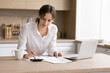 © fizkes - Young woman using calculator notebook to review documents at kitchen