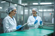 © Vadym - Two women in hygiene uniforms and gloves check product quality on a factory conveyor belt. One records data on a clipboard while the other speaks. Clean facility operation.