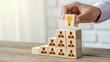 © Ham - Hand placing wooden block with lightbulb icon atop human figure pyramid on wooden table in innovative team leadership concept.