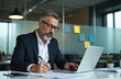 © Maryna - Mature businessman intently reviews paperwork at his desk in modern office. He signs documents and analyzes financial reports on a laptop. Focused pro works diligently with business strategy.