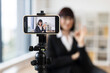 © sofiko14 - Caucasian woman wearing formal attire recording video blog using smartphone on tripod in modern office setting. Concept themes include business networking, educational content creation.