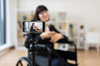 © sofiko14 - Caucasian woman seated in wheelchair recording blog on smartphone about business topics. Bright room with tripod and background bookshelf provide professional and creative environment for filming.