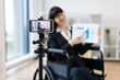© Sofiia Shunkina - Caucasian woman in wheelchair blogs on camera about business using charts and graphs. Indoor setting with focus on professional blogging and business concepts.