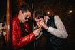 © qunica.com - Two young men lean close in a dim, vintage space, sharing a phone screen and laughing. One wears a red leather jacket, the other a white shirt and vest.