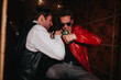 © qunica.com - Two men share a candid moment during a night out, one in a red leather jacket and sunglasses. They laugh and pose with a bottle, creating a lively social vibe.