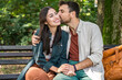 © Srdjan - Young couple cuddling on a park bench, smiling and leaning in for a kiss, sharing an affectionate embrace in sunny green nature casual outfits, warm connection, romantic date outdoors