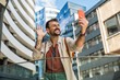 © Srdjan - Young traveler laughing while taking a selfie in a modern business district cheerful millennial man with backpack capturing urban architecture and city vibes on smartphone during a sunny day