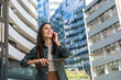 © Srdjan - Smiling young woman on phone call in modern business district cheerful traveler with backpack, chatting on smartphone amid sleek urban architecture and reflective office towers on a sunny day