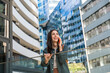 © Srdjan - Smiling young woman on phone call in modern business district cheerful traveler with backpack, chatting on smartphone amid sleek urban architecture and reflective office towers on a sunny day