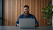 © Roshan - Smiling Black Man Working on Laptop at Desk in Modern Office with Wood Paneling and Plant for Business, Technology, and Professional Concepts