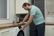 © Anna Baranova - Man doing laundry at home in modern kitchen. Person loading washing machine and pouring detergent. Concept of household chores, domestic life, and home cleaning routine