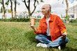 © Bliss - Bald woman enjoying her coffee while sitting on grass in orange denim jacket outdoors
