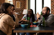 © DC Studio - Young adults pause from board game to listen intently as white man shares an engaging story. Diverse friends gathered around the table with alcoholic beverage and bowl of chips, hanging out indoors.
