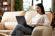 © DC Studio - Asian businesswoman sitting comfortably on couch and researching company data progress on laptop. Professional female freelancer enjoying productive work session in cozy home office.