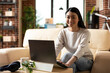 © DC Studio - Cheerful female entrepreneur sitting confidently on sofa with laptop in home office. Smiling businesswoman working remotely, ready to review online articles to boost company progress.