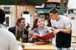 © JackF - On open area cafe terrace, guy waiter communicates with girl and her boyfriend customers, answer questions about composition of dish of day. Client consult with waiter about vegan health menu