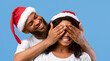 © Prostock-studio - Excited african american man closing wife's eyes, surprised young woman spreading arms to sides and smiling, couple wearing Sants hats, standing together over yellow studio background