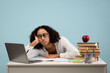 © Prostock-studio - Tired black female student studies at desk surrounded by books and a laptop. She rests her head on her hand, showing signs of exhaustion, with a blue background emphasizing her struggle.