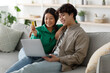 © Prostock-studio - A joyful Asian couple sits on a comfortable sofa, using a laptop to shop online. The woman holds a credit card, and they share a close moment while enjoying their time together.