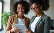 © yuan - Two happy busy professional business women, diverse female employees workers talking using digital tablet device working together in partnership while standing at work in office. Candid photo.